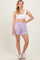 woman wearing a white crop top and lavender shorts, standing with one hand on her hip, smiling at the camera