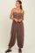 woman wearing a brown sleeveless jumpsuit with button details and flowy pants, standing against a light background