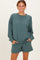 woman wearing a teal oversized sweatshirt and matching shorts, smiling with hands in pockets
