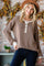 woman wearing a brown henley sweater with decorative buttons and cream detailing, smiling with a hat in a cozy setting