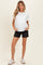 woman wearing a white short sleeve maternity top and black shorts, posing with hands in pockets