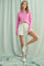 woman wearing a pink button-up long sleeve cardigan with white shorts and white sneakers standing against a light backdrop