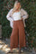 woman wearing a white tank top, oversized striped shirt, and brown wide leg pants standing in front of greenery