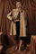 woman wearing a tan oversized trench coat over a brown cropped top and black capri pants, posing against a draped background