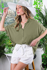 woman wearing an olive green oversized button-up shirt and white shorts, standing in a bright greenery-filled setting