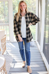 woman wearing a black and tan plaid oversized shirt, white top, and blue jeans, standing in a light-filled room