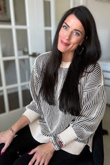 woman wearing a cream and black striped oversized sweater, sitting and smiling, with long dark hair and accessories