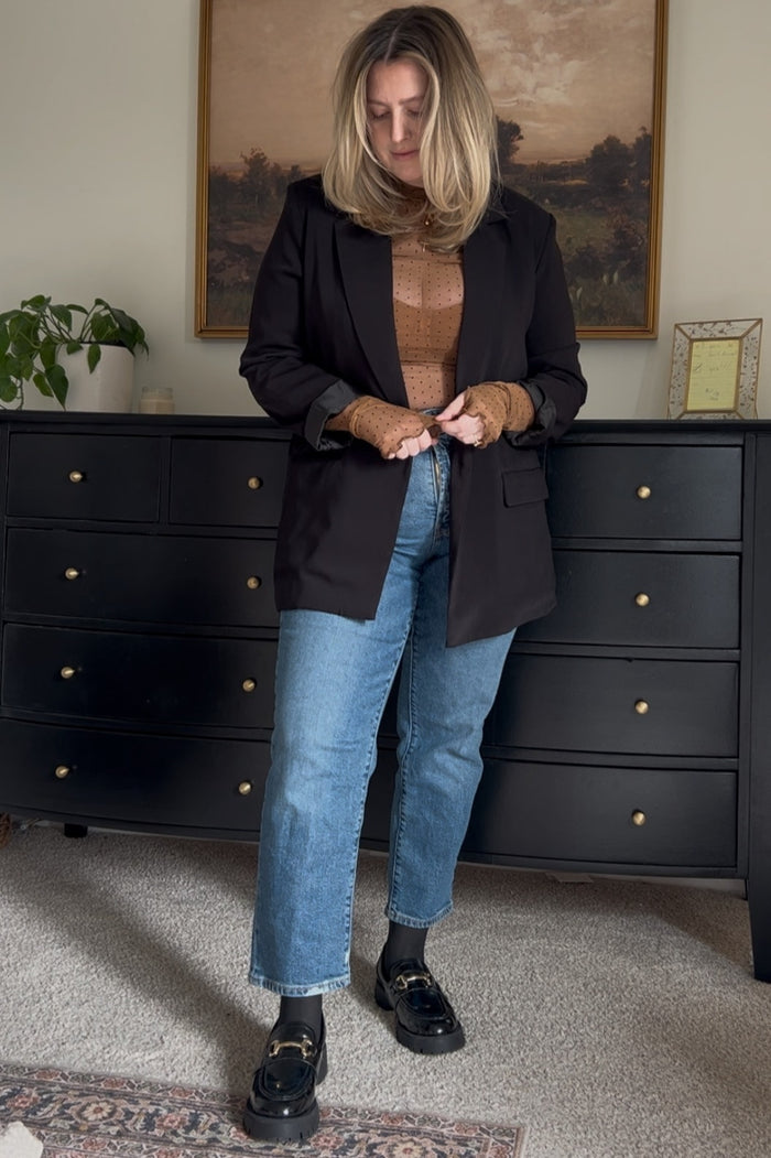 woman wearing a black blazer over a brown knit top and blue denim jeans, styled with black loafers and wavy hair
