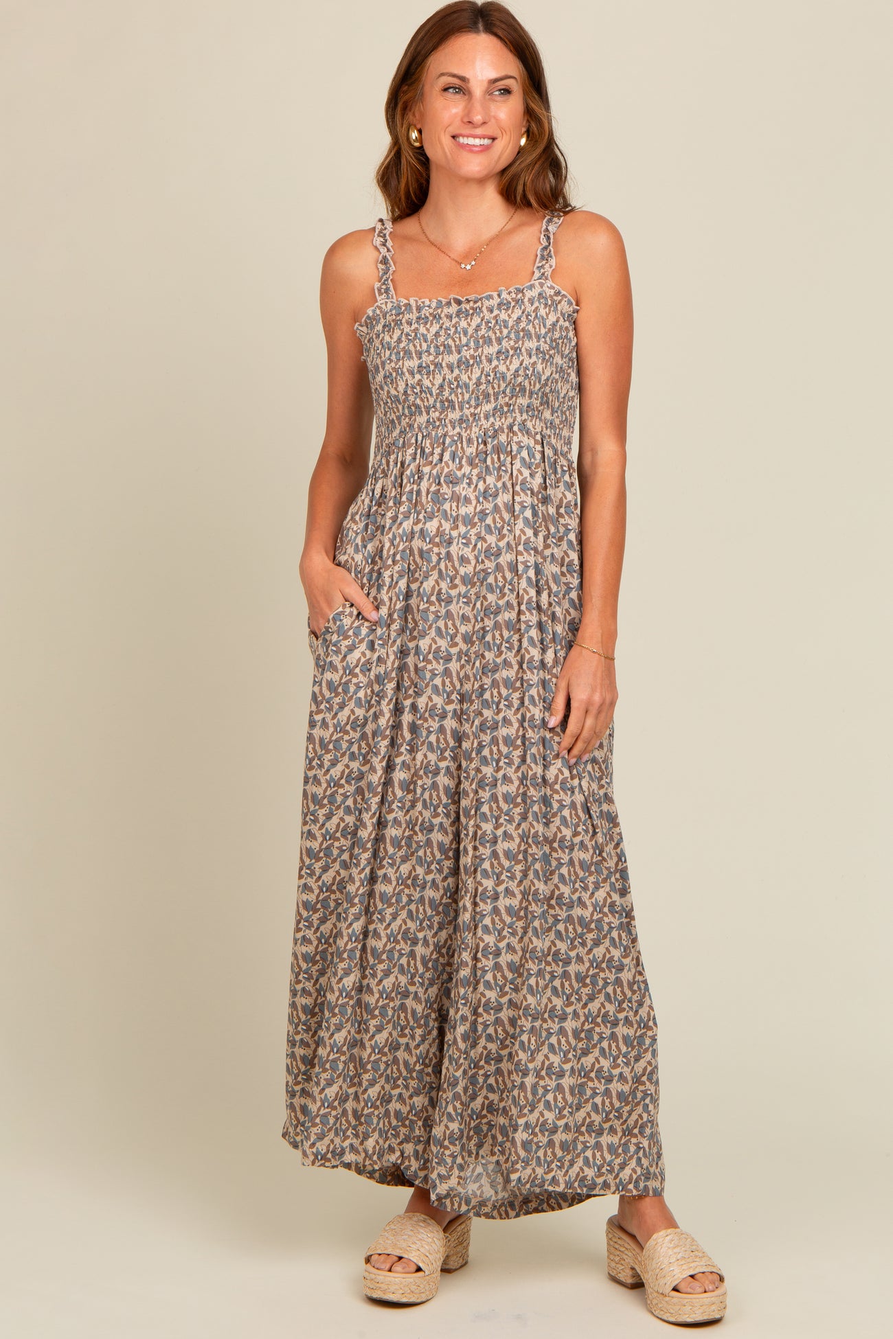 woman wearing a brown floral maxi dress with adjustable straps and pockets, standing and smiling