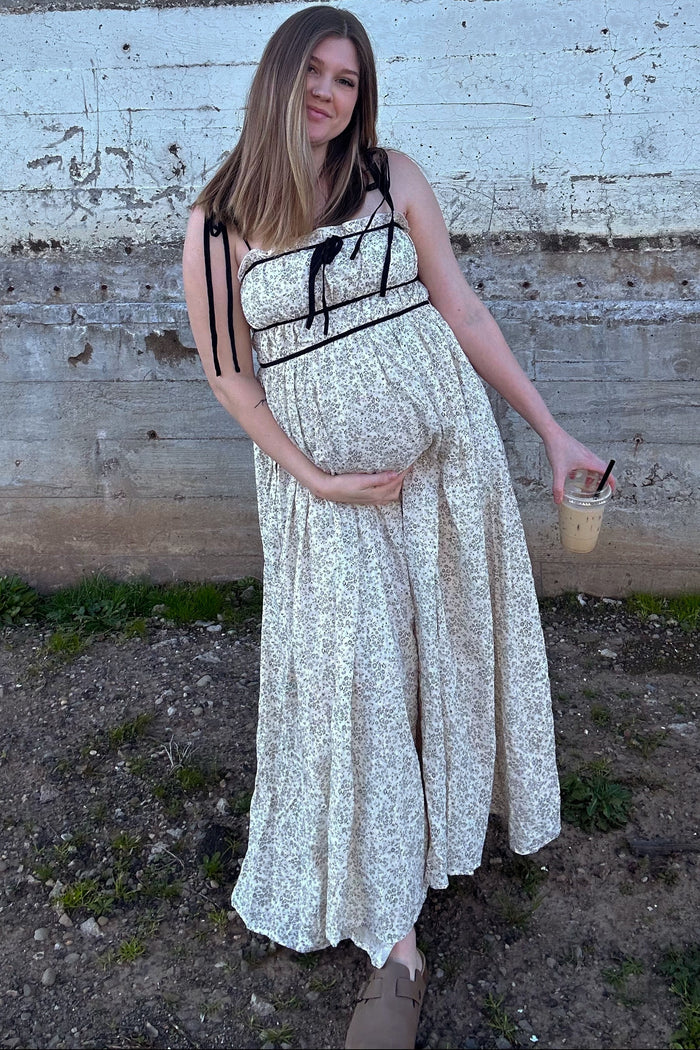 woman wearing a floral print maternity maxi dress and holding her baby bump while holding an iced drink