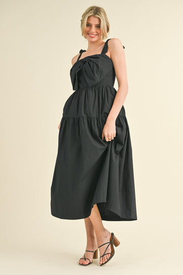 woman wearing a black tiered midi dress with bow straps and a flowing silhouette, standing on a light background