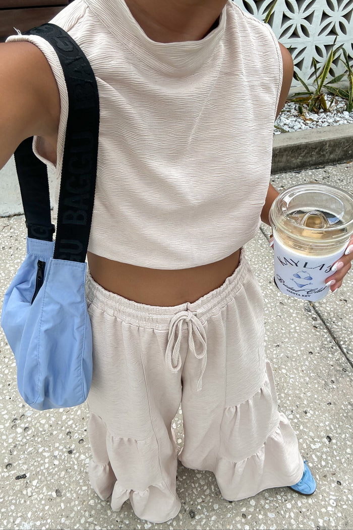woman wearing a cropped beige top with a cowl neck and matching tiered wide-leg pants, holding a drink and a blue bag