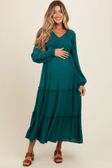 woman wearing a teal maternity maxi dress with long sleeves and tiered ruffle detail, holding her baby bump