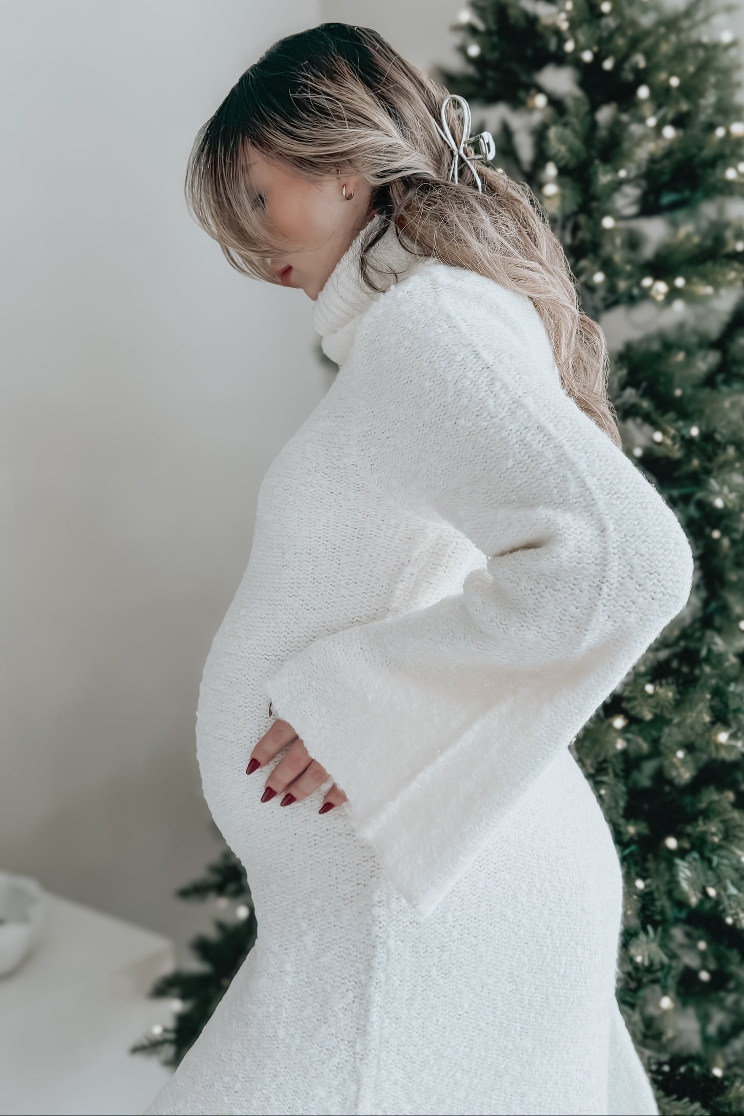 woman wearing a white maternity sweater dress, holding her baby bump, standing next to a decorated Christmas tree