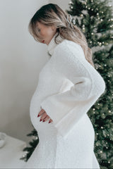 woman wearing a white maternity sweater dress, holding her baby bump, standing next to a decorated Christmas tree