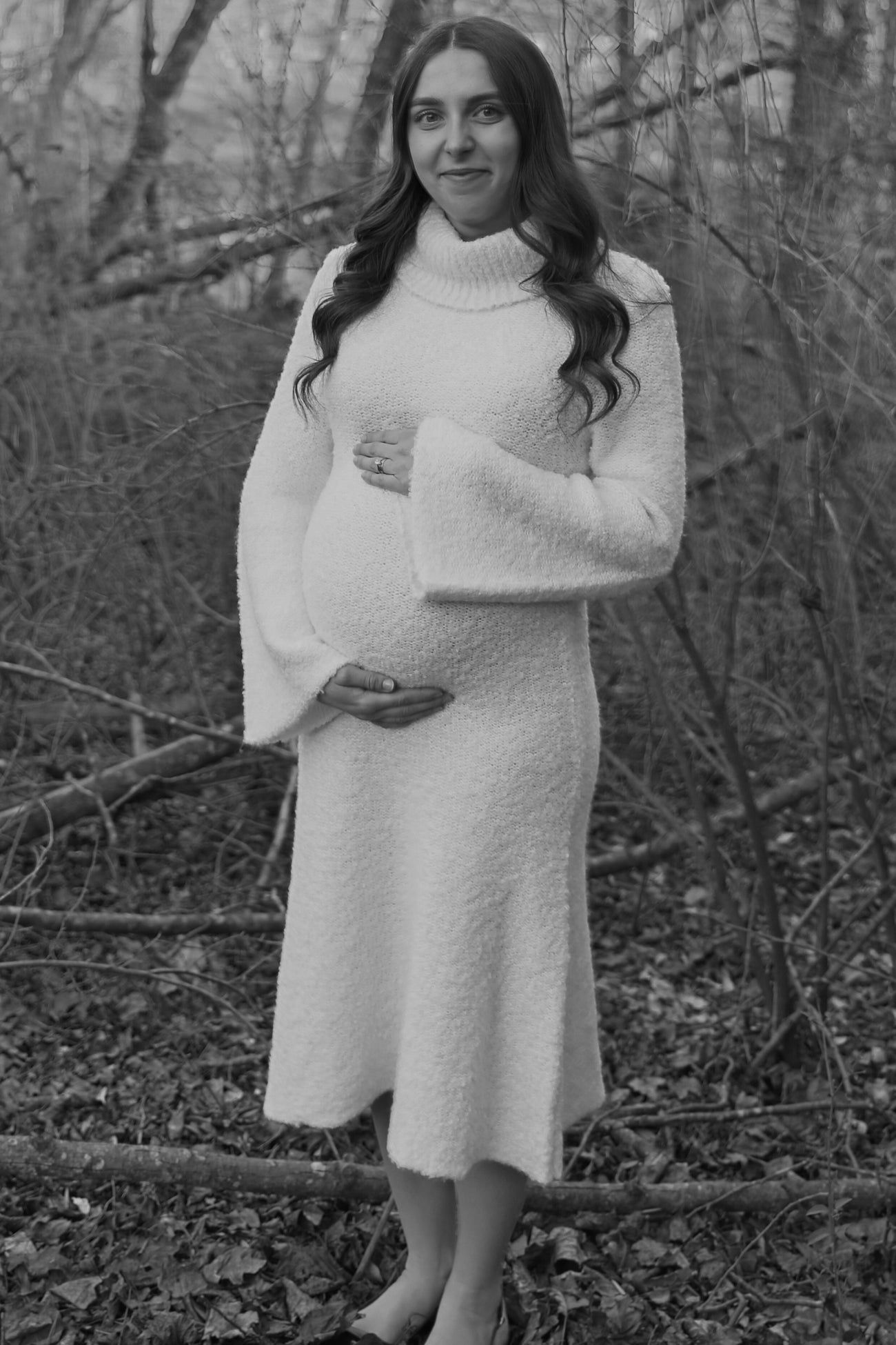 woman wearing a cozy white maternity dress with flared sleeves, gently cradling her baby bump in a natural setting