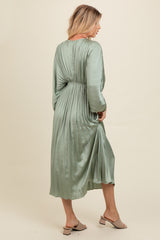 woman wearing a green pleated midi dress with long sleeves and a cinched waist, standing sideways