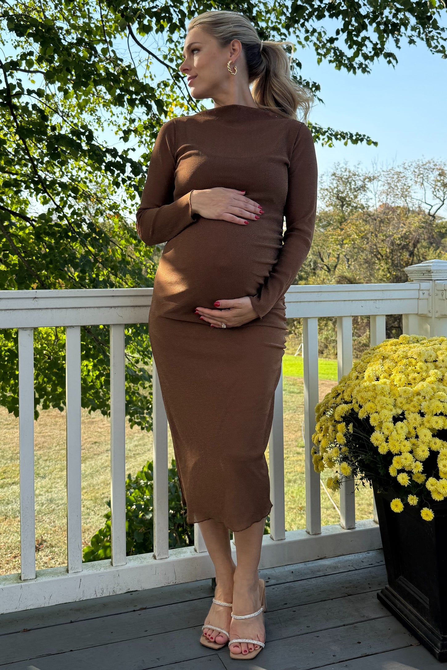 woman wearing a brown long sleeve maternity dress and holding her baby bump while standing on a porch
