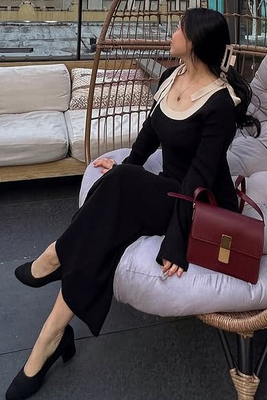 woman sitting on a chair wearing a black long sleeve dress with a cream collar and holding a burgundy handbag