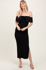 woman wearing a black off-the-shoulder midi dress with a bow detail and side slit, standing with hands on her hips