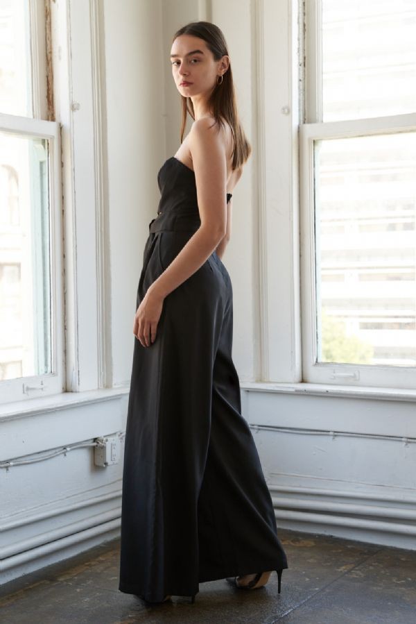 woman wearing a black strapless wide-leg jumpsuit, standing near a window with a modern aesthetic