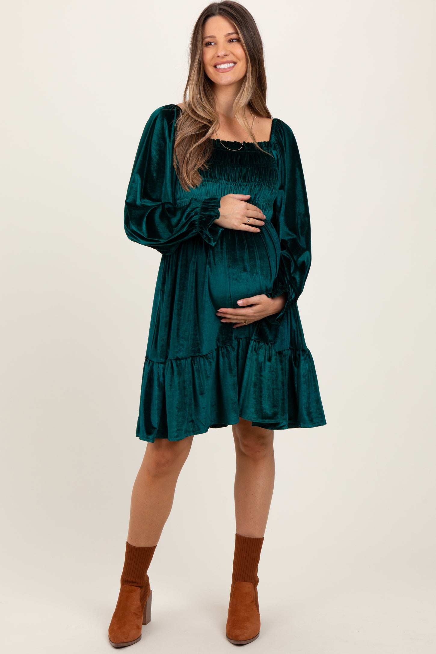 woman wearing a dark green velvet maternity dress with long sleeves, smocked bodice, and tiered skirt, holding her baby bump