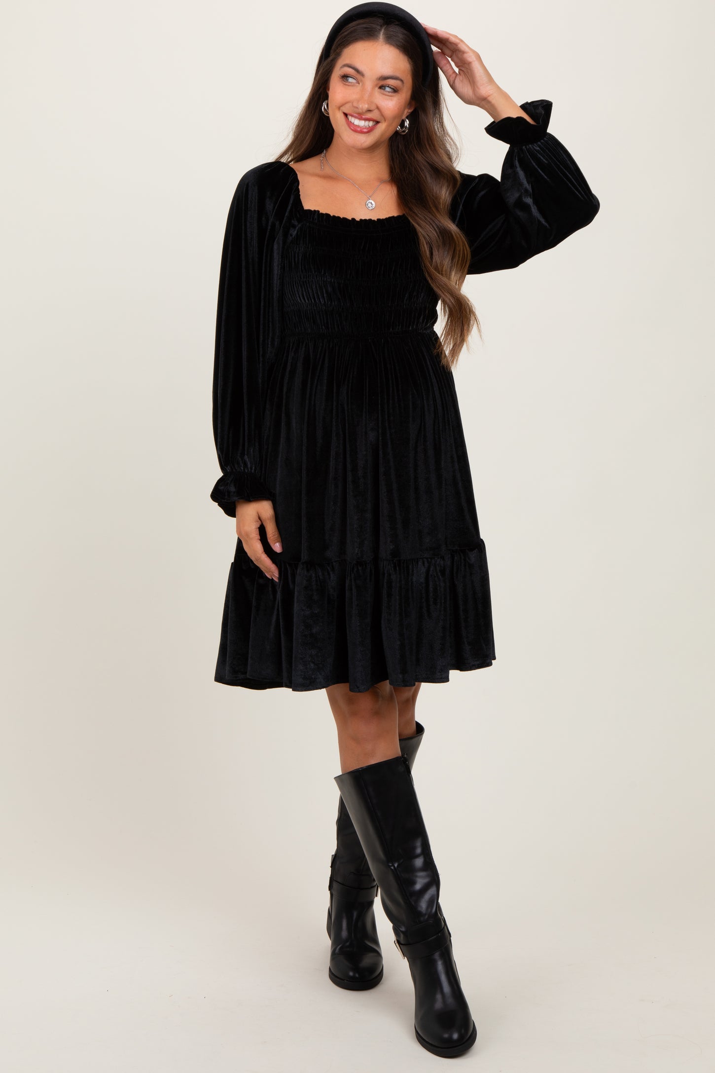 woman wearing a black velvet maternity dress with smocked bodice and puff sleeves, standing with hand in hair
