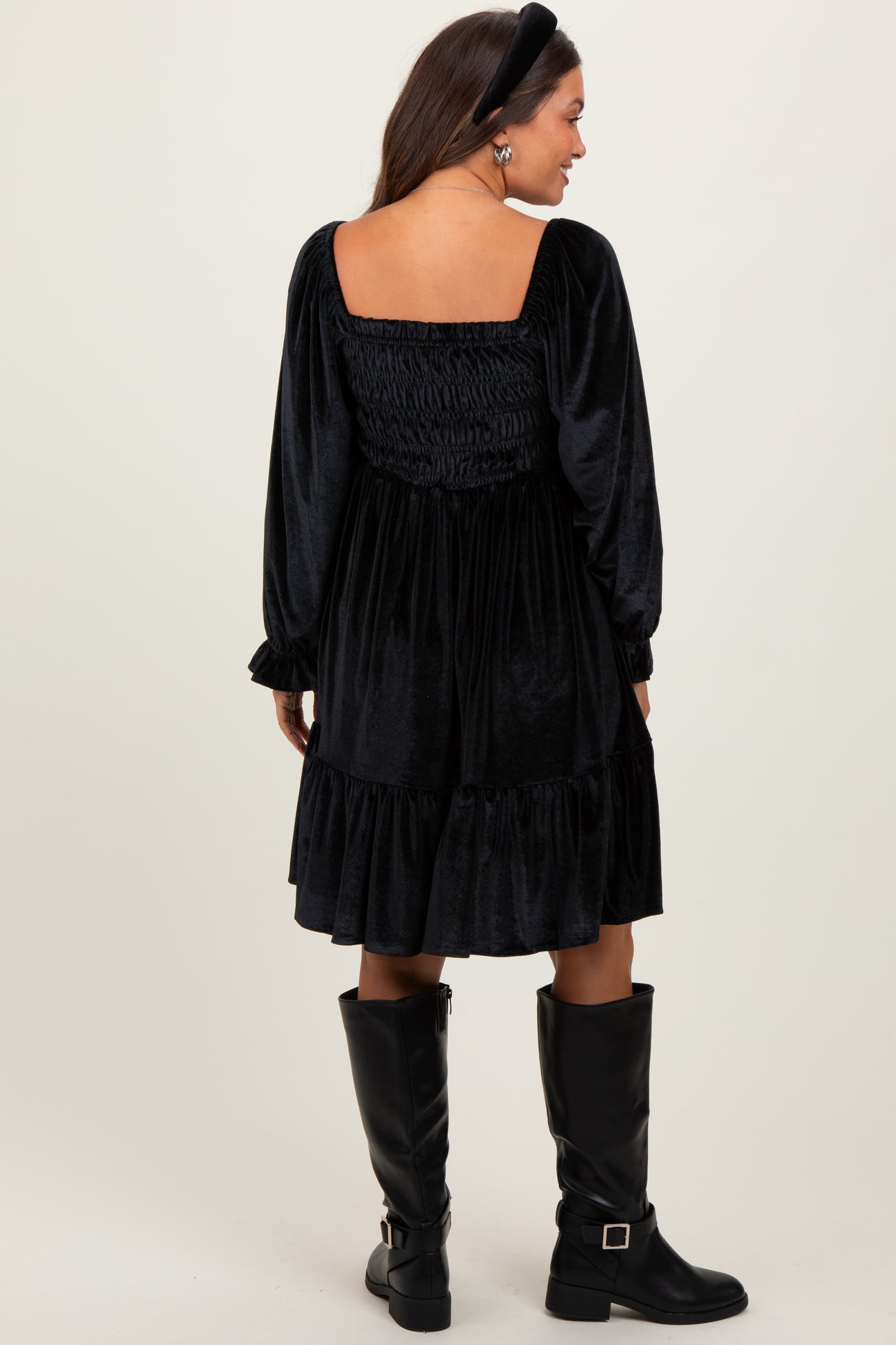 woman wearing a black velvet maternity dress with a smocked back and long sleeves, paired with knee-high black boots