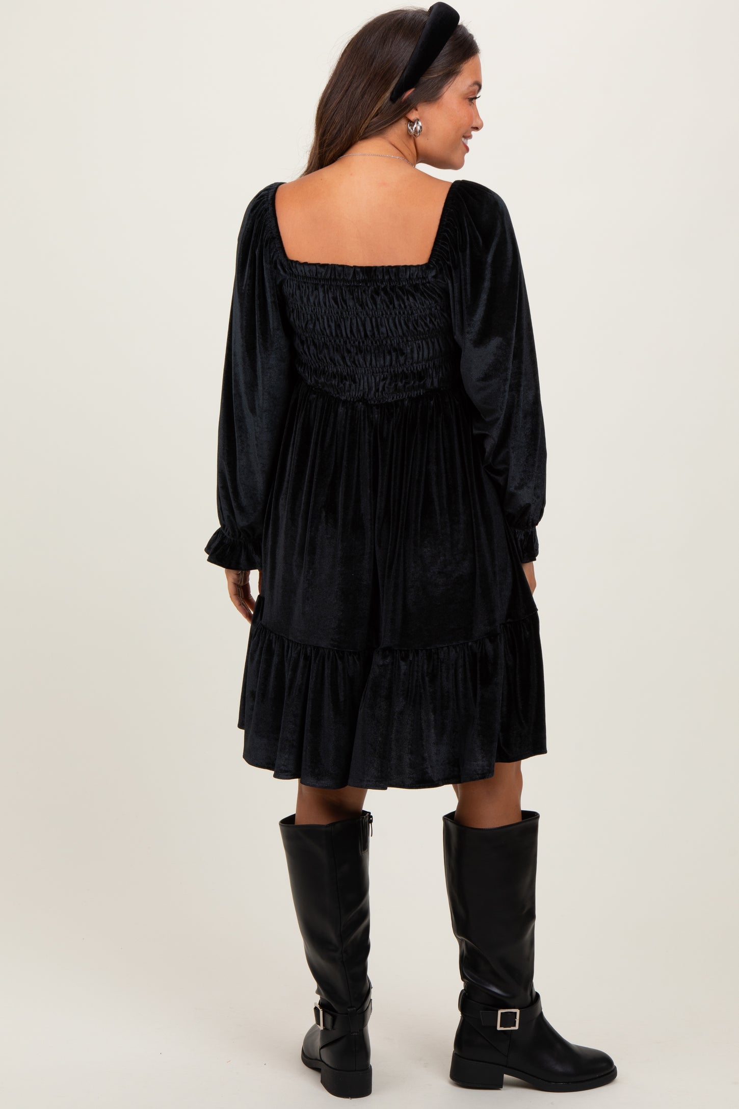 woman wearing a black velvet maternity dress with a smocked back and long sleeves, paired with knee-high black boots