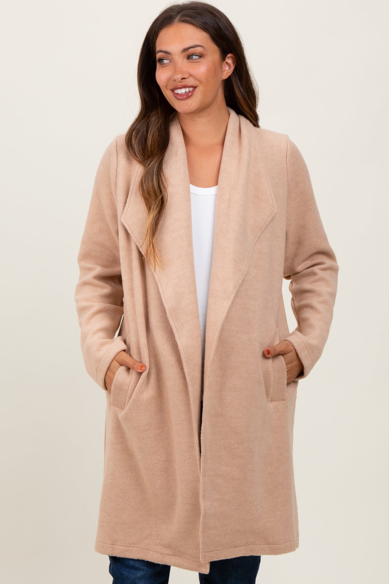woman wearing a beige maternity wrap coat with pockets, styled open over a white top and denim jeans