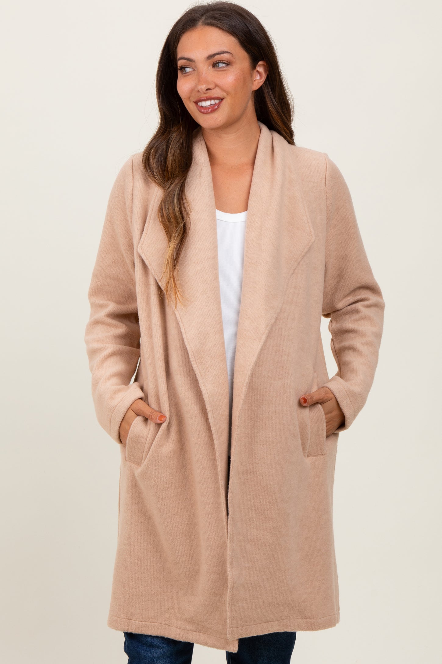 woman wearing a beige maternity wrap coat with pockets, styled open over a white top and denim jeans