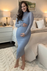 woman wearing a light blue long sleeve maternity dress, cradling her baby bump in a cozy bedroom setting
