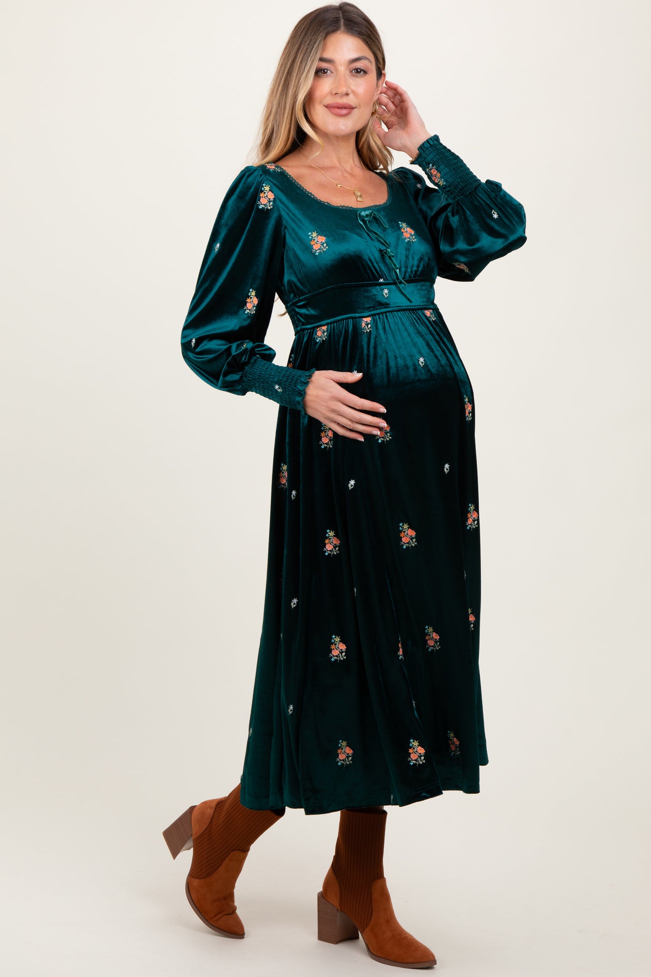 woman wearing a deep green velvet maternity dress with floral embroidery and long sleeves, visible baby bump