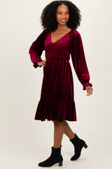 woman wearing a burgundy velvet dress with long sleeves, smocked bodice, and a ruffled hem, posing with a smile
