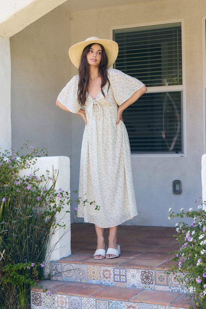 woman wearing a floral midi dress with short sleeves and a tie front, standing on a tiled porch with a wide-brimmed hat