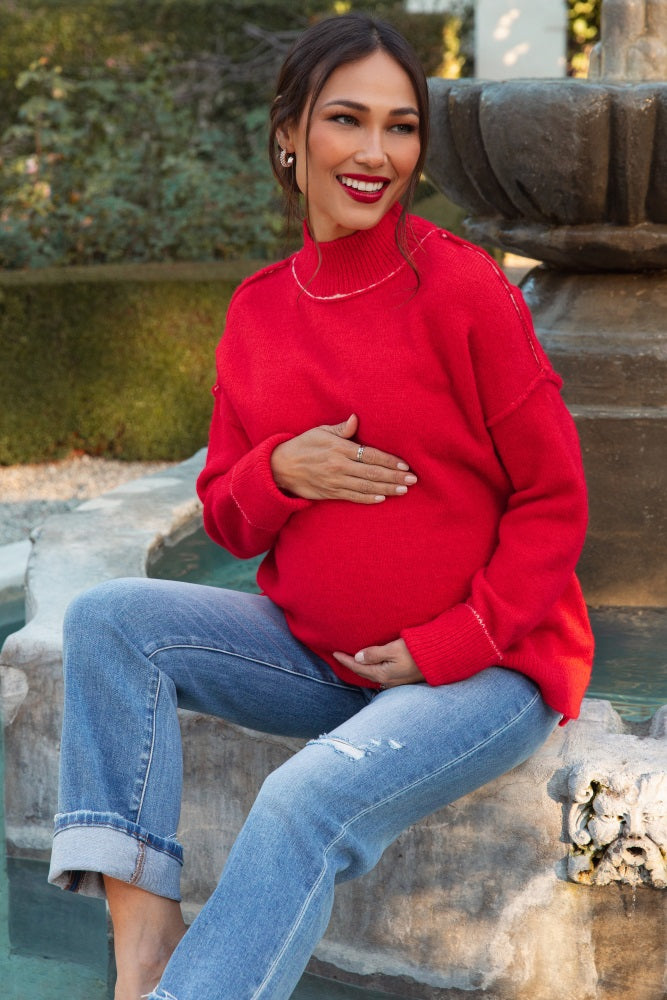 woman wearing a red oversized maternity sweater and distressed blue jeans, holding her baby bump by a fountain