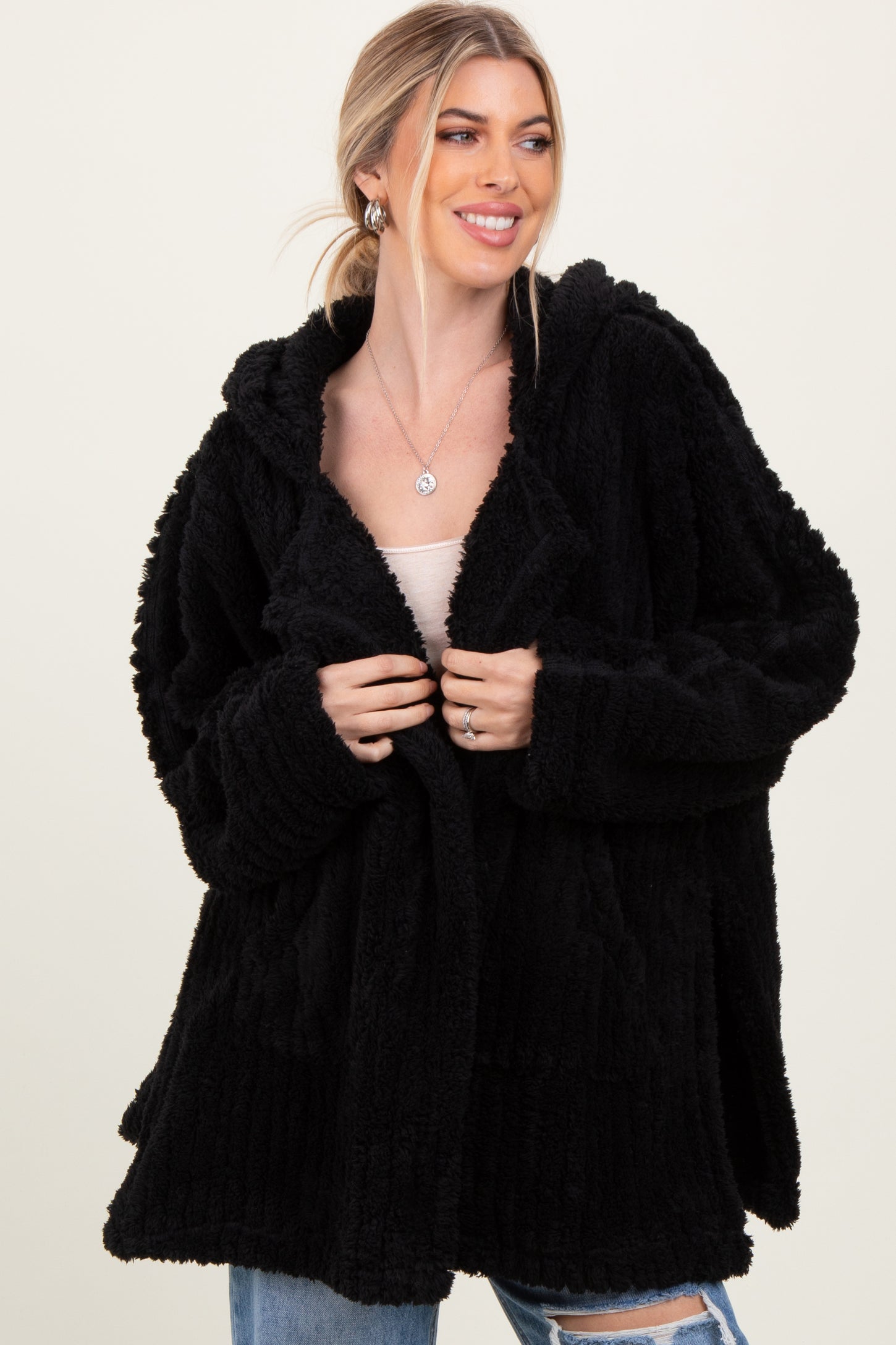 woman wearing a black plush hooded cardigan, smiling and holding it open over a beige top