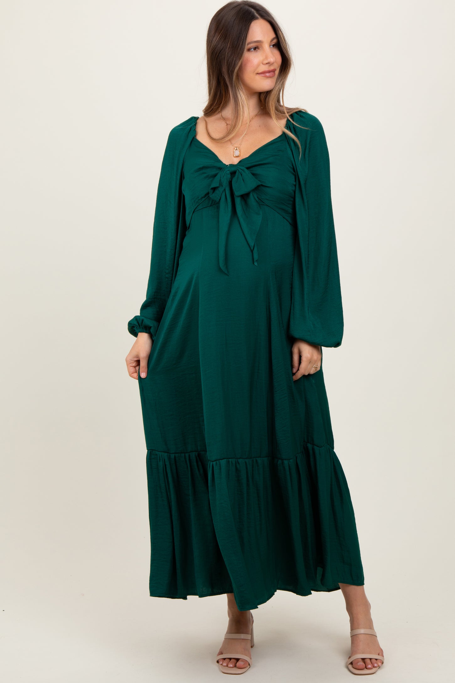 woman wearing a dark green maternity dress with a tie front and long sleeves, featuring a tiered skirt and a visible baby bump