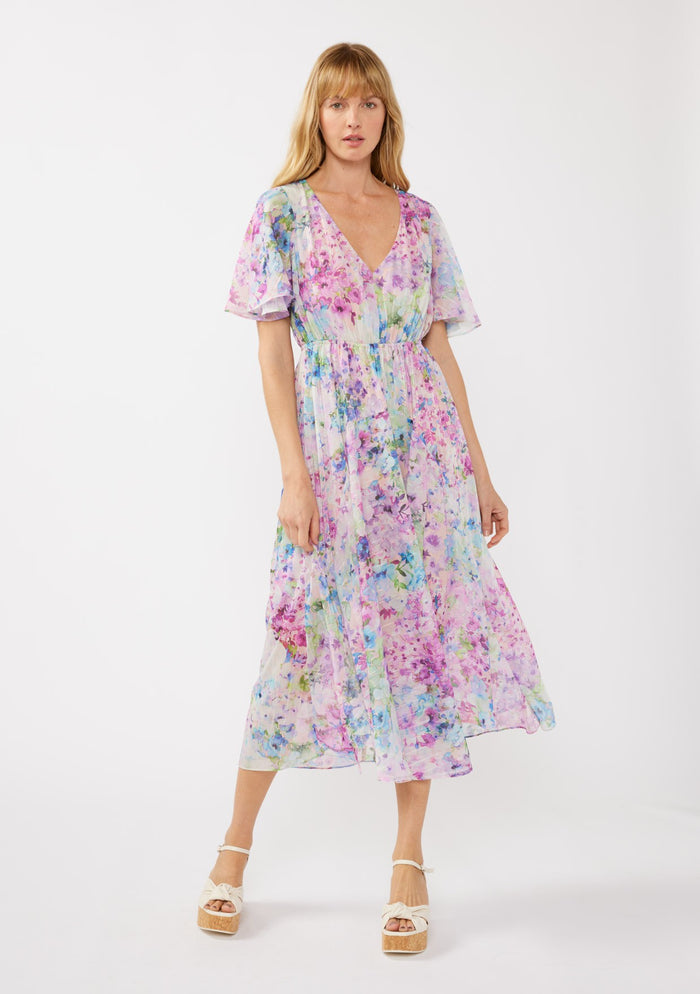 woman wearing a long, flowing floral maxi dress in pastel shades of purple, pink, and blue with short sleeves