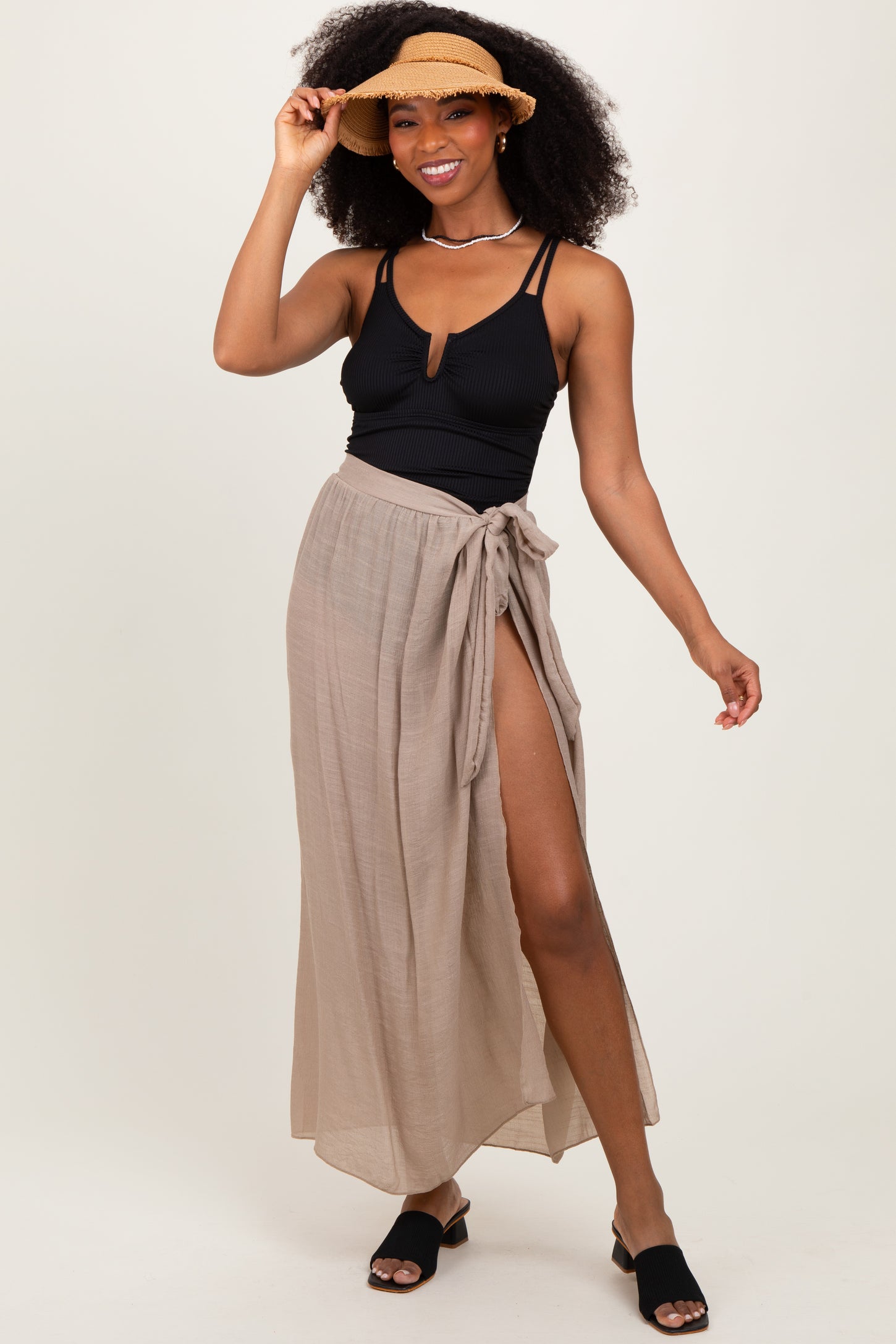 woman wearing a black ribbed swimsuit top and a tan wrap maxi skirt, accessorized with a straw hat and slip-on sandals