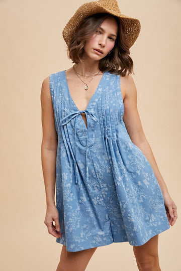 woman wearing a light blue floral summer dress with a tie detail and pleated design, accessorized with a straw hat