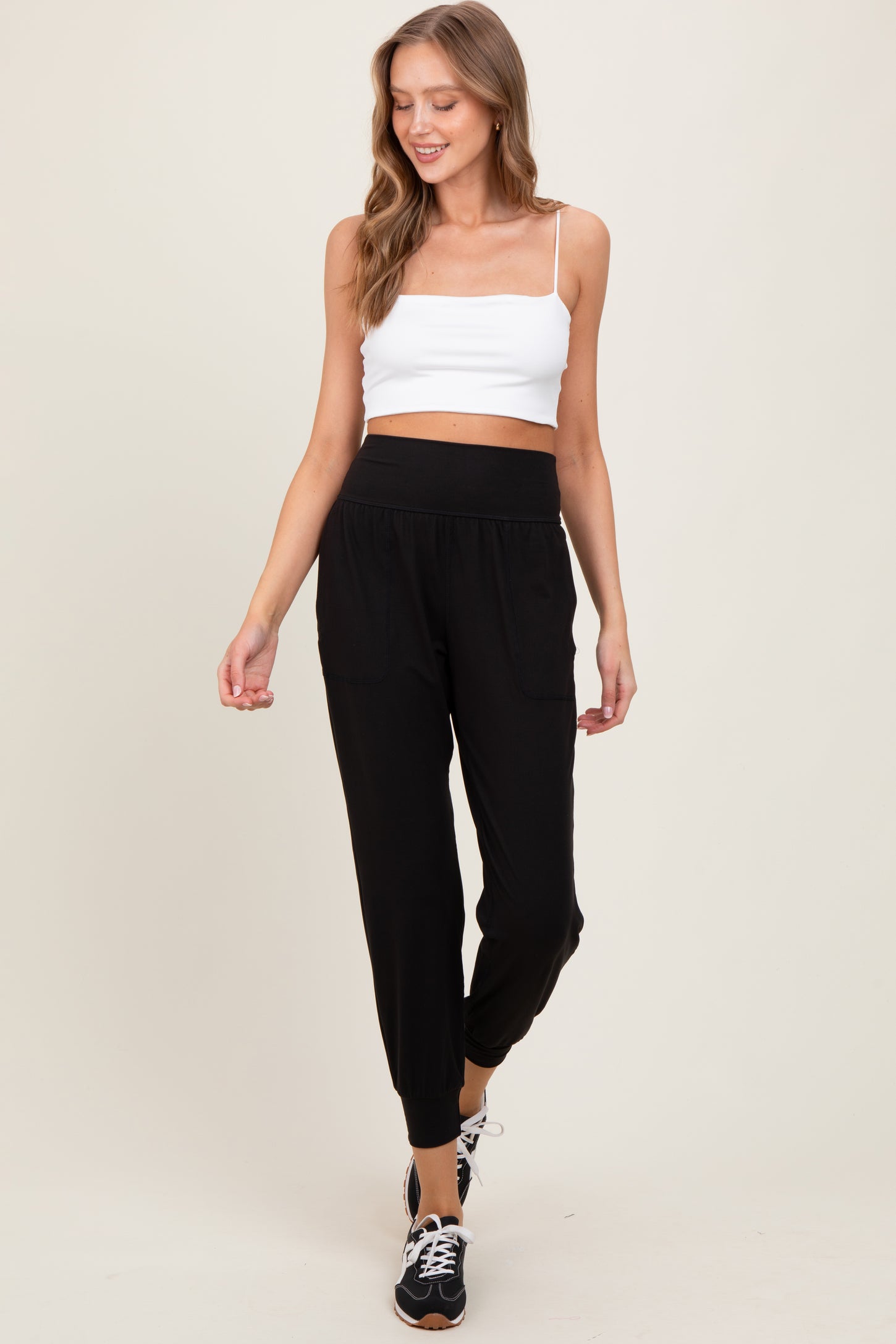 woman wearing a white cropped tank top and black jogger pants with pockets, smiling and walking against a neutral background