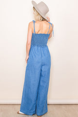 woman wearing a back view of a denim jumpsuit with smocked bodice and wide-leg pants, accessorized with a wide-brim hat