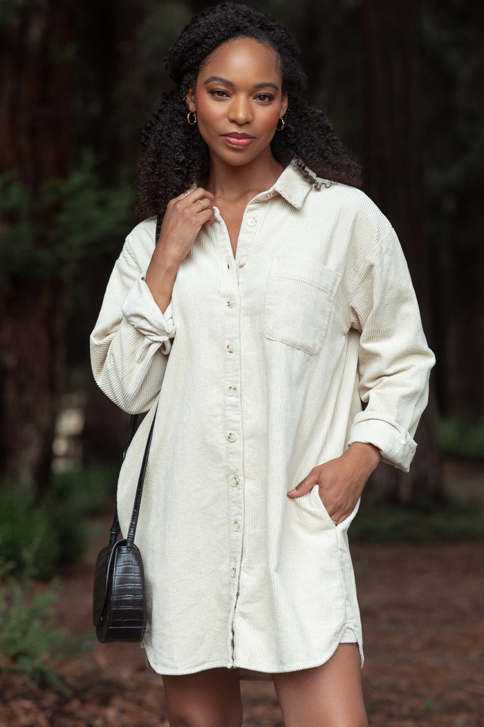 woman wearing a beige corduroy shirt dress with a button front, long sleeves, and a side pocket, standing outdoors