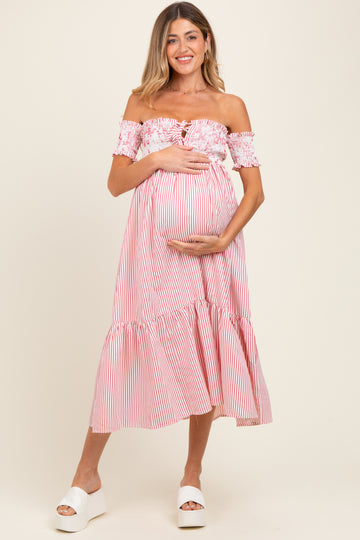 woman wearing a pink and white striped off-the-shoulder maternity dress, holding her baby bump with a smile