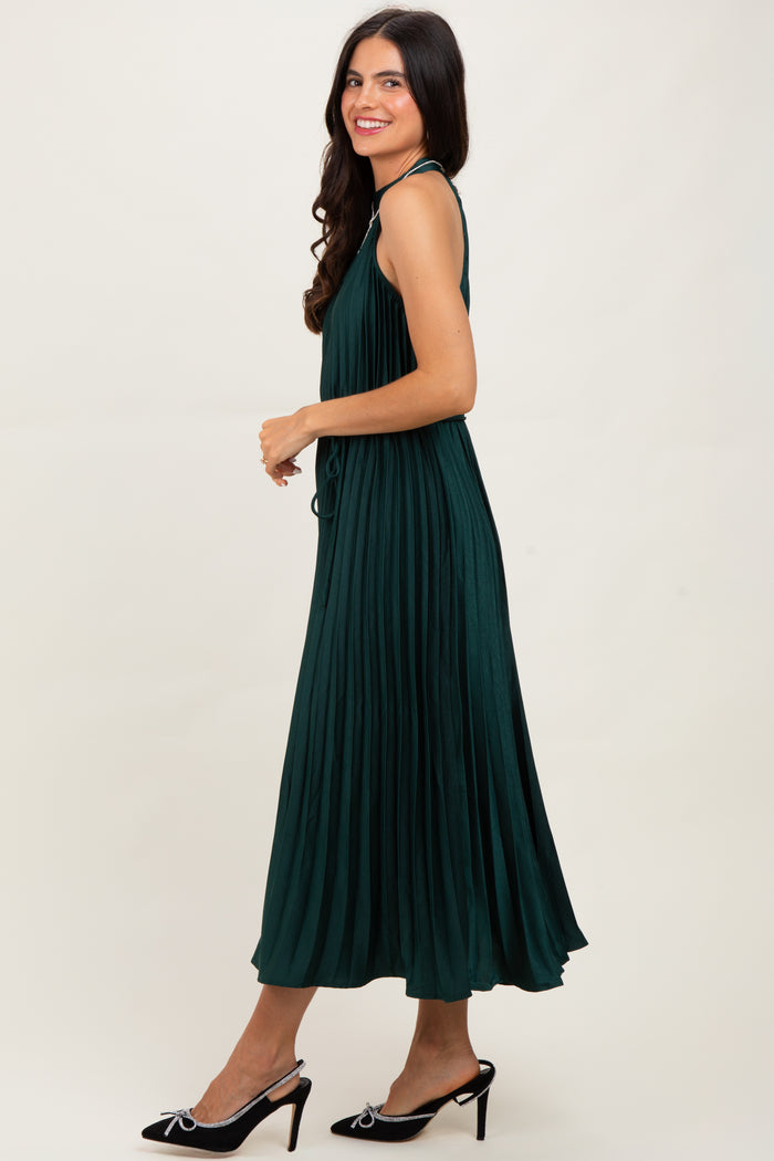 woman wearing a dark green pleated maxi dress with straps and high heels, standing sideways against a neutral background
