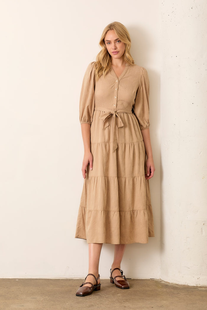 woman wearing a tan tiered midi dress with puff sleeves and a tie waist, standing against a wall