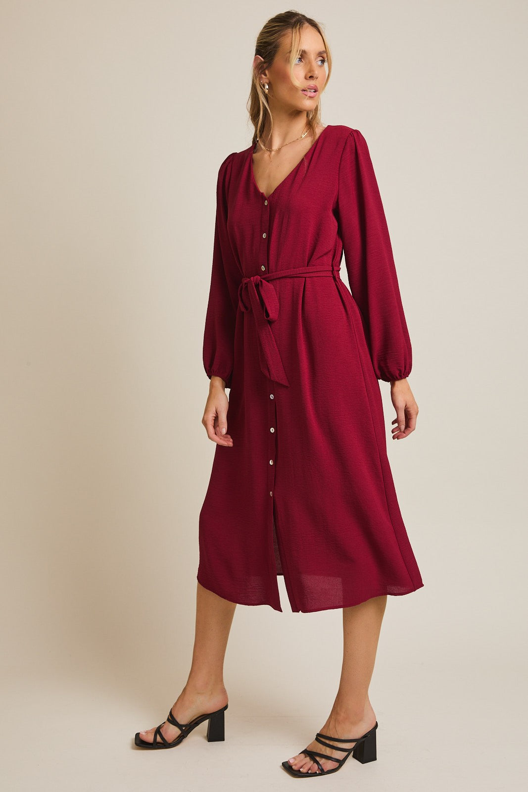woman wearing a burgundy midi dress with long sleeves, button front, and a tie belt, styled with black strappy heels