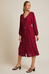 woman wearing a burgundy midi dress with long sleeves, button front, and a tie belt, styled with black strappy heels