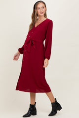 woman wearing a burgundy long sleeve maternity dress with a tie waist and button details, showcasing her baby bump